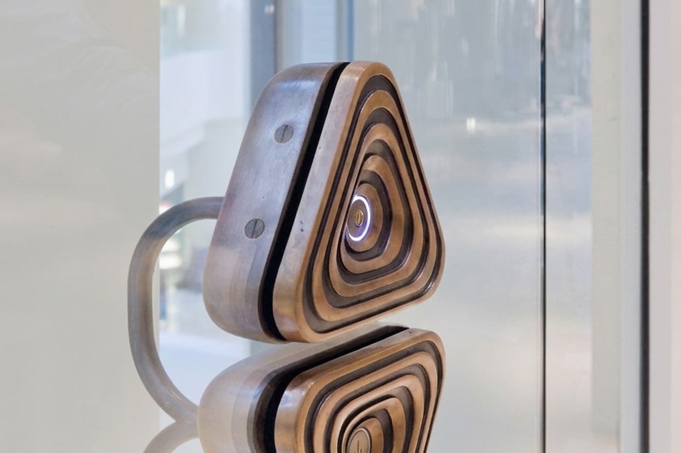 A bronze lift landing call button on a glass lobby wall, its call ring illuminated, reflected in the polished surface below.