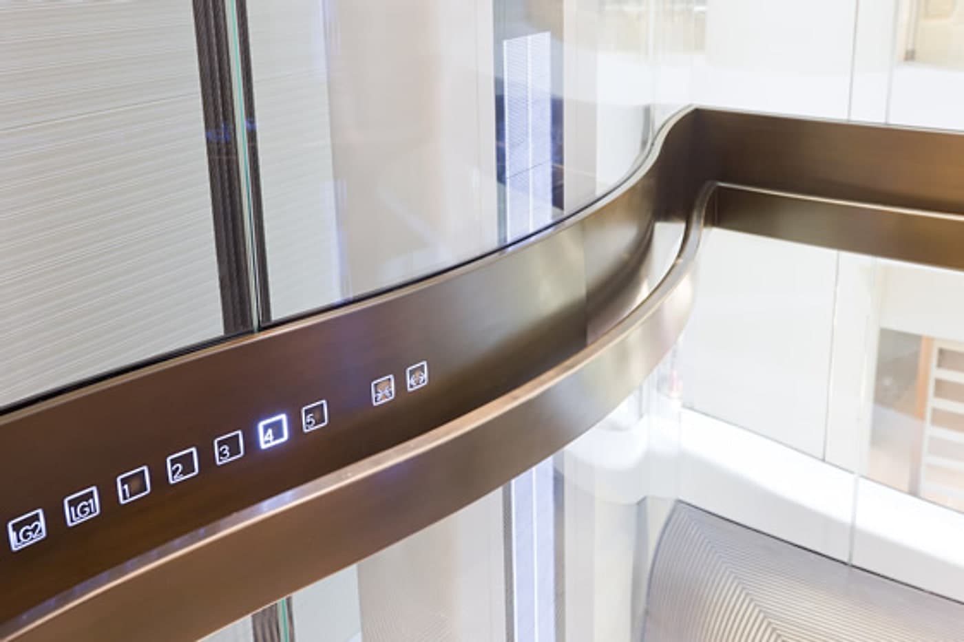 A lift landing call panel set into the curved bronze balustrade of a shopping centre atrium, glass lift shafts behind.