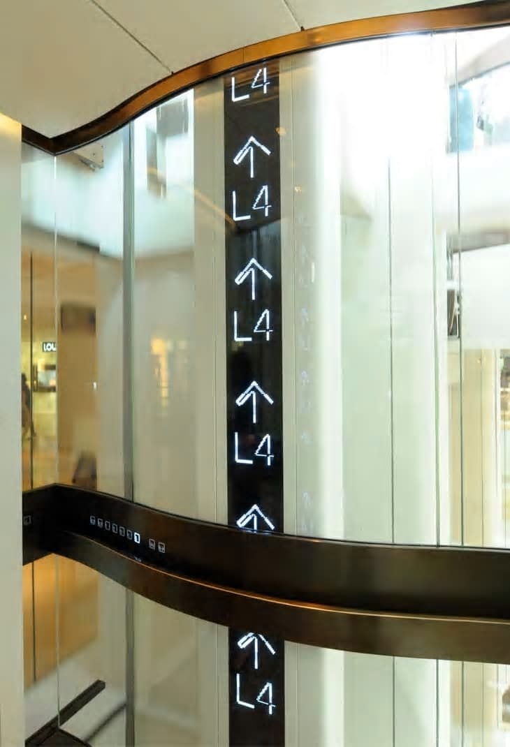 Glass lift shafts in a shopping centre lobby, position indicators showing level four, curved bronze balustrade trim.
