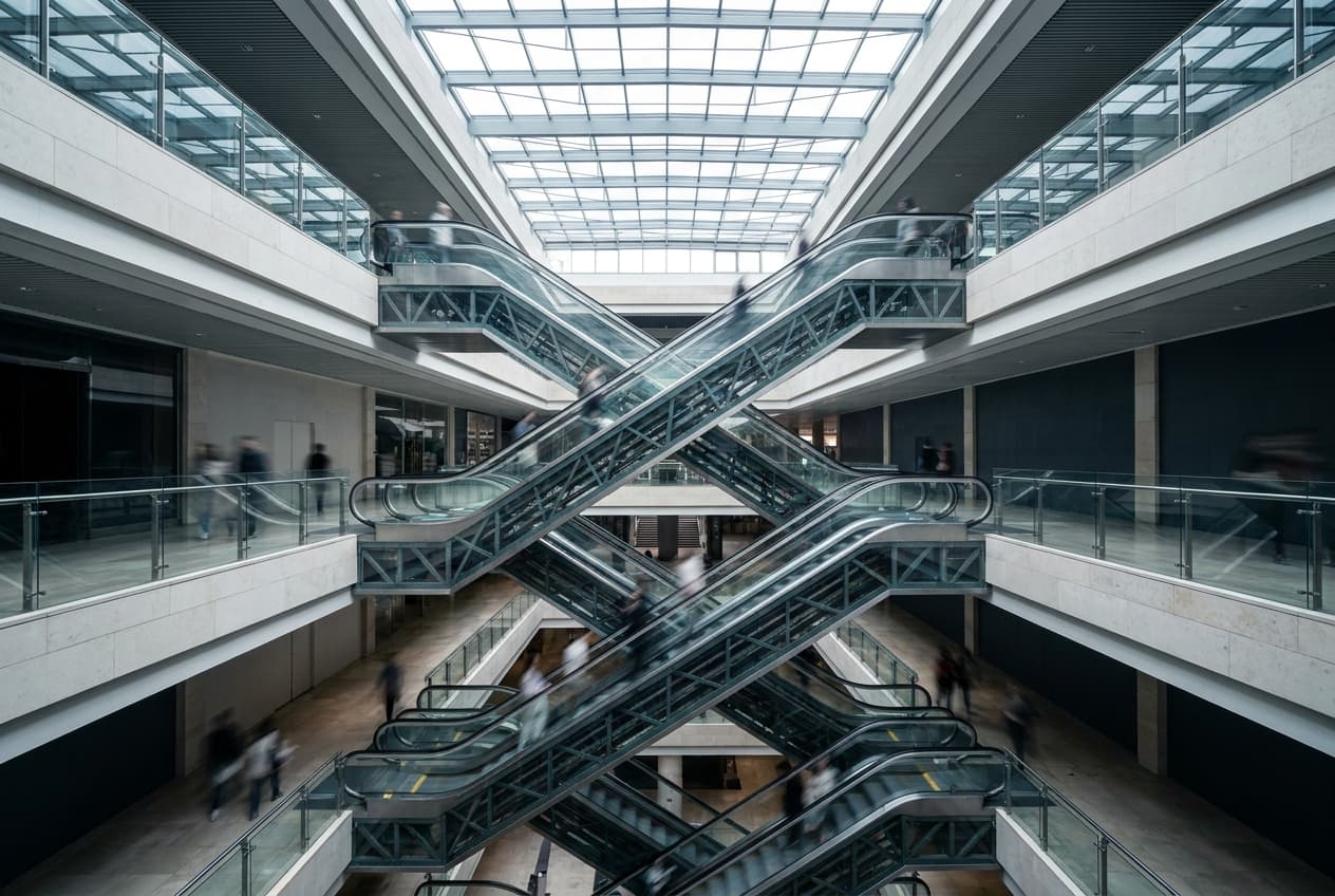Escalators criss-crossing a full-height shopping centre void through three retail levels, shoppers moving on each floor.