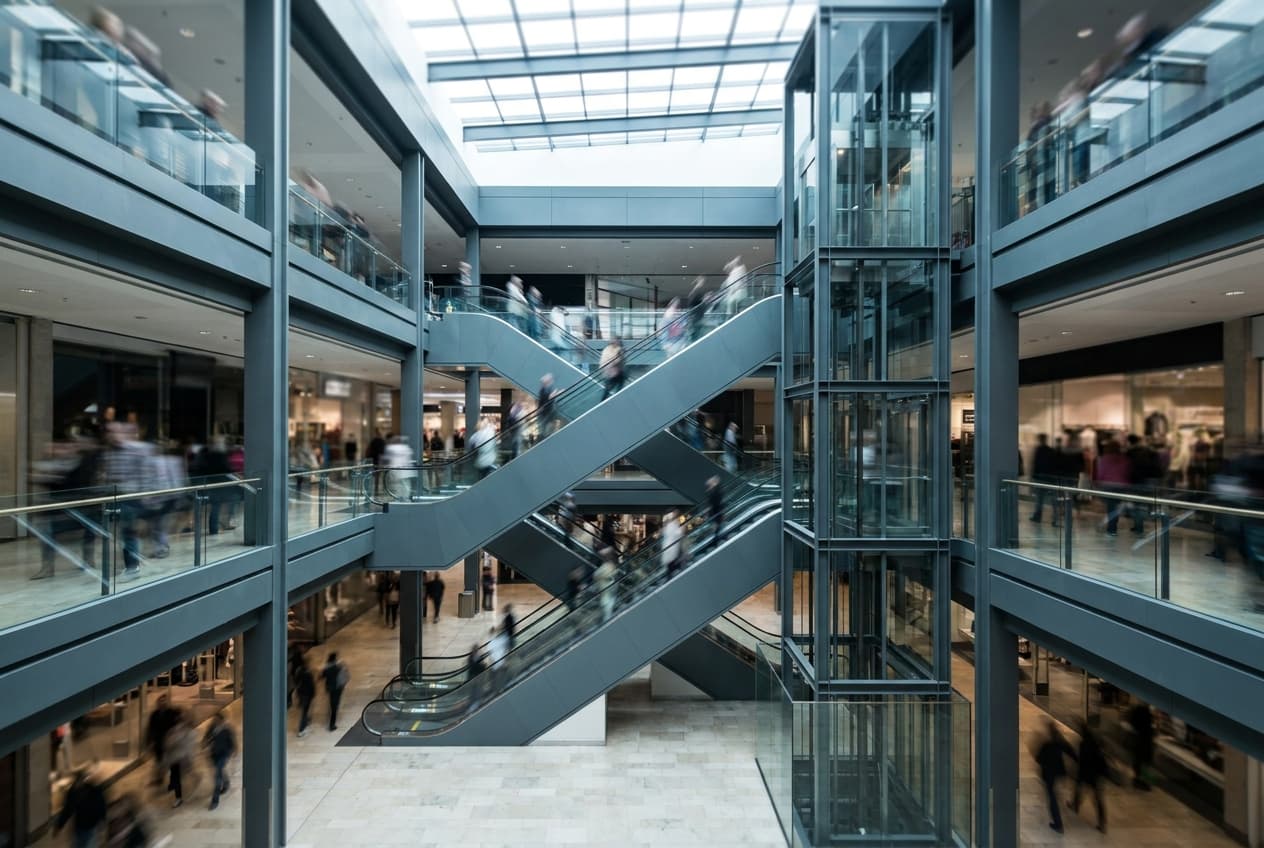A three-level shopping centre void with escalators crossing between the floors and a glass lift beside them, shoppers moving on every level.
